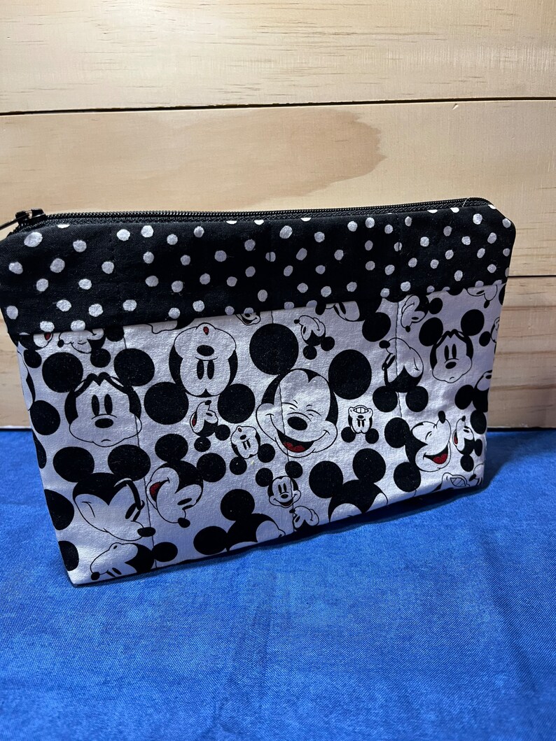 Puede incluir: Una bolsa con cremallera negra y blanca con un estampado de Mickey Mouse. La bolsa tiene un ribete de tela de lunares negros y blancos.