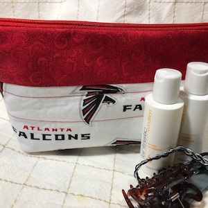 May include: Red and white zippered pouch with the Atlanta Falcons logo. The pouch is sitting on a white surface with two bottles of hair care products and hair clips.