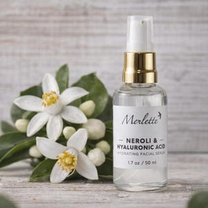 May include: A clear glass bottle of Merlette Neroli & Hyaluronic Acid Hydrating Facial Serum, 1.7 oz / 50 ml, with a gold-colored cap. The bottle is next to white flowers with yellow centers and green leaves.