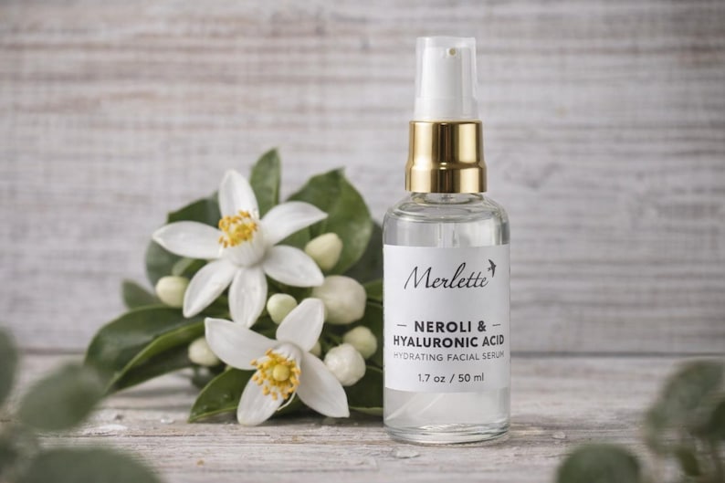 May include: A clear glass bottle of Merlette Neroli & Hyaluronic Acid Hydrating Facial Serum, 1.7 oz / 50 ml, with a gold-colored cap. The bottle is next to white flowers with yellow centers and green leaves.