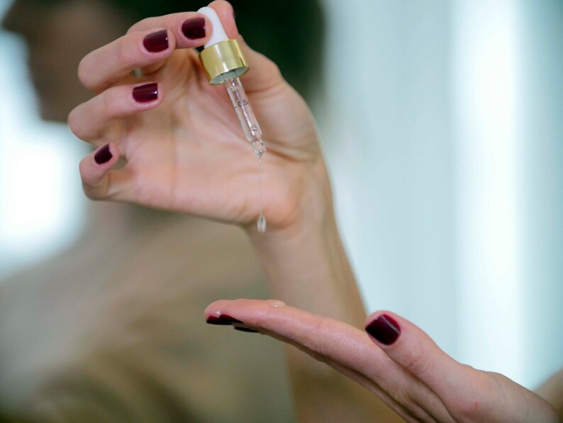 May include: A clear glass dropper bottle with a gold cap dispenses a liquid onto a person's hand. The person's nails are painted a dark red color. The background is blurred, suggesting a focus on skincare.