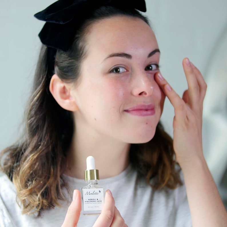 May include: A woman with a black bow in her hair applies serum from a small glass bottle. The bottle, labeled "Merlette Neroli & Hyaluronic Acid," has a white dropper and gold accents. The serum bottle is 0.5 fl oz / 15 ml.