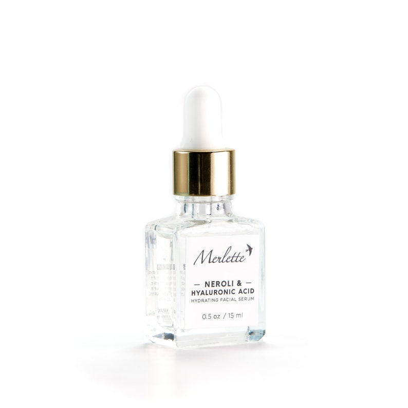 May include: A clear glass bottle of Merlette Neroli & Hyaluronic Acid Hydrating Facial Serum. The bottle has a white dropper top with a gold collar and a white label with black text. The bottle contains 0.5 oz / 15 ml of serum.