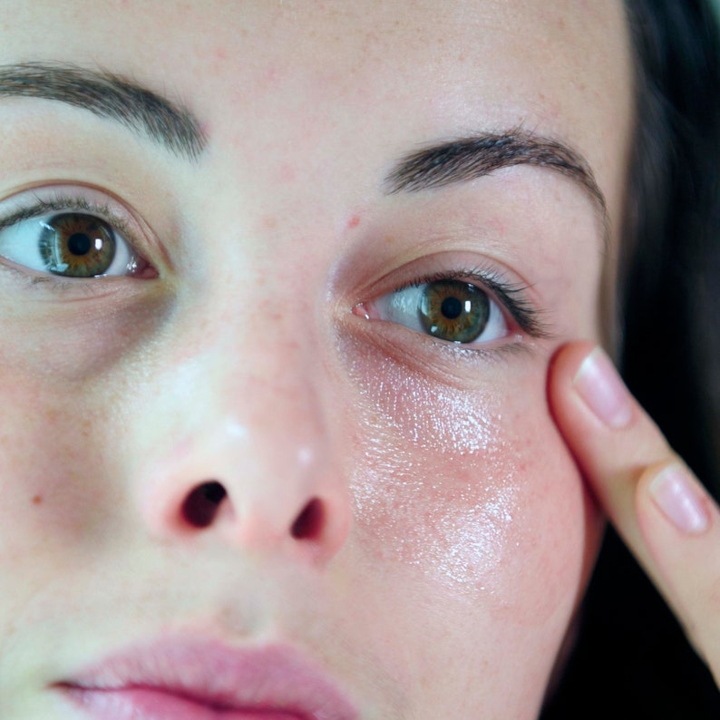 May include: Close-up of a person's face with green eyes and dark eyebrows. The skin appears to be glowing, with a finger gently touching the area under the eye. The focus is on the eye and the surrounding skin.