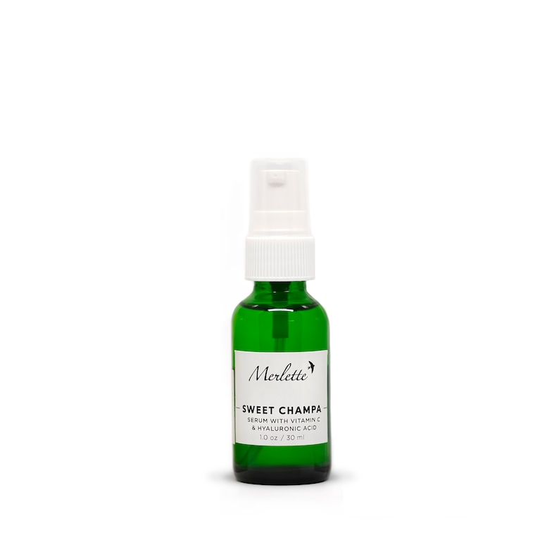 Peut inclure: Une bouteille en verre vert avec un vaporisateur blanc. La bouteille porte une &eacute;tiquette blanche avec le texte "Merlette SWEET CHAMPA SERUM WITH VITAMIN C & HYALURONIC ACID 30 ml". Sur fond blanc.
