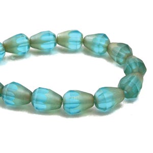 May include: A strand of aqua and pale green glass beads. Each bead is teardrop-shaped with a faceted surface and a light blue floral design at the top. The beads are strung together, creating a curved line against a white background.