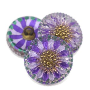 May include: Three decorative buttons with a floral design. The buttons feature a purple and clear petal design with gold-coloured centres. The edges are trimmed with green and clear beads. One button has a purple and green striped design.