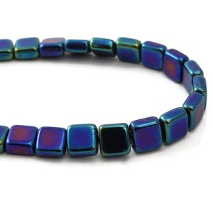 May include: Close-up of a strand of iridescent square beads. The beads shift in color from blue to purple and green, with a glossy finish. The beads are approximately 0.25 inches square, strung together.