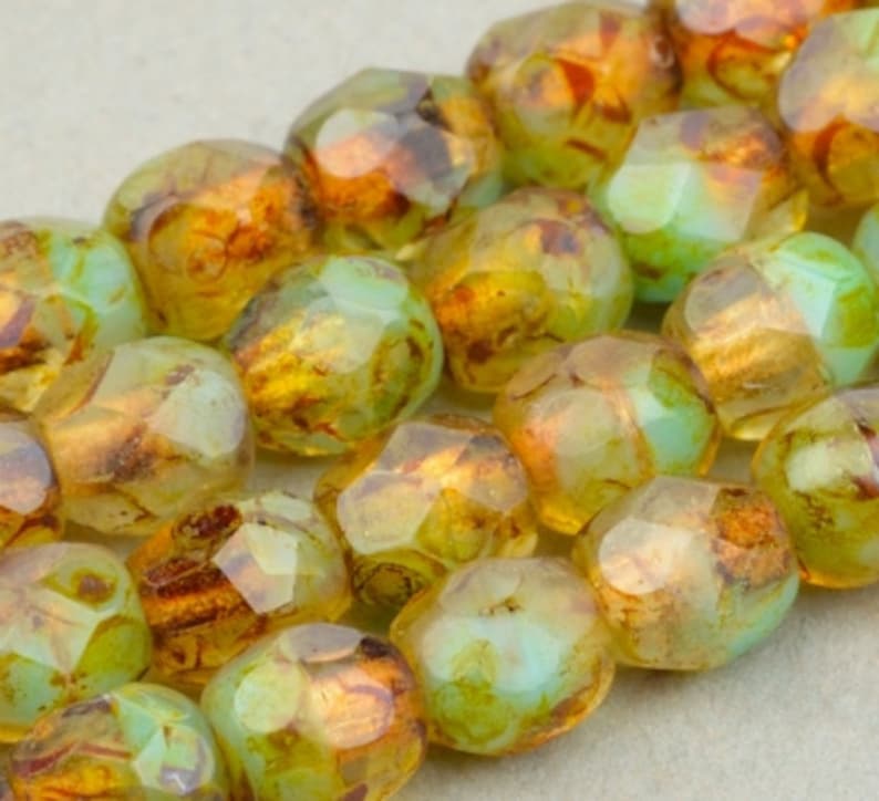May include: A strand of faceted glass beads in shades of green and gold with an iridescent finish. The beads have a unique, textured surface.