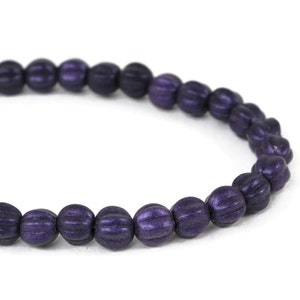 May include: A close-up shot of a string of dark purple beads. Each bead has a ribbed texture, resembling a small pumpkin. The beads are strung together, forming a curved line against a white backdrop. The beads are a deep, saturated purple colour.