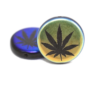 May include: Two iridescent, round beads with a black cannabis leaf design. One bead is in focus, displaying a gold and green hue, while the other is out of focus, showing a blue color. The beads have a hole for stringing.