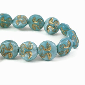 May include: A strand of turquoise-colored beads with a gold-colored, stylized design. The beads are round and have a slightly irregular shape. The gold design appears to be a repeating pattern. The beads are strung together, suggesting they are for jewelry making.