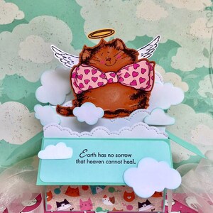 May include: A pop-up card with a brown cat wearing a pink bow tie and angel wings. The cat is surrounded by white clouds and the card reads "Earth has no sorrow that heaven cannot heal."