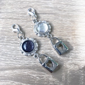 Op de afbeelding: Twee zilveren kettingen met een hartvormige slot hanger en een ronde hanger met een glinsterend blauwe en een heldere glazen kern.