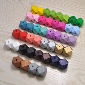 Puede incluir: Un conjunto de 15 cuentas de madera en varios colores, cada cuenta tiene forma de diamante con un agujero en el centro. Las cuentas están dispuestas en línea de izquierda a derecha, con los colores pasando del blanco al dorado.