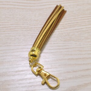 Puede incluir: Un llavero con borla dorada y cierre de mosquetón. La borla está hecha de tiras superpuestas de cuero sintético marrón y dorado. Los herrajes metálicos son de color dorado brillante.