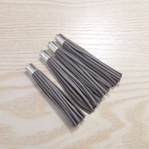 Könnte beinhalten: Fünf graue Wildlederquasten mit silberfarbenen Metallkappen werden auf einer hellen Holzmaserungsoberfläche präsentiert. Die Quasten sind diagonal angeordnet und unterstreichen ihre weiche Textur. Diese dekorativen Quasten eignen sich für verschiedene Bastelprojekte.