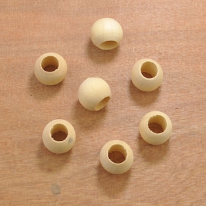 May include: Seven unfinished wooden beads with a natural, light-coloured finish. Each bead is spherical with a large, centred hole. The beads are arranged in a circular pattern on a light brown wooden surface.