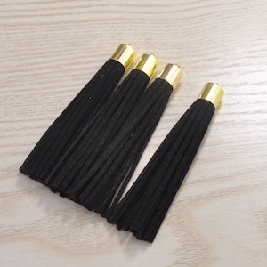 Puede incluir: Cuatro borlas de gamuza negra con tapas de metal dorado. Las borlas están dispuestas diagonalmente sobre una superficie clara con vetas de madera. Cada borla está hecha de muchos hilos finos y deshilachados, creando una apariencia suave y texturizada. Estas borlas decorativas son adecuadas para varios proyectos de manualidades.