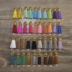 Pode incluir: Um conjunto de 39 borlas de camurça coloridas com pontas de metal dourado. As borlas estão dispostas em filas sobre uma superfície de madeira.