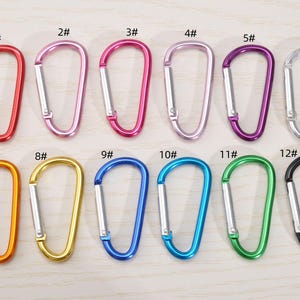May include: A selection of twelve carabiner clips in a range of colours: red, pink, purple, silver, orange, gold, blue, green, and black. Each clip has a silver gate and is numbered from 1 to 12.