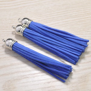May include: Three blue suede tassels with silver-colored metal caps and loops. The tassels are arranged on a light wood-grain surface. These decorative tassels are suitable for jewelry making, crafting, or embellishing various items.