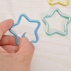 May include: A hand holding a blue star-shaped carabiner clip. Several other star-shaped clips in various colors, including green and gold, are visible in the background. The clips have a metallic, iridescent finish.