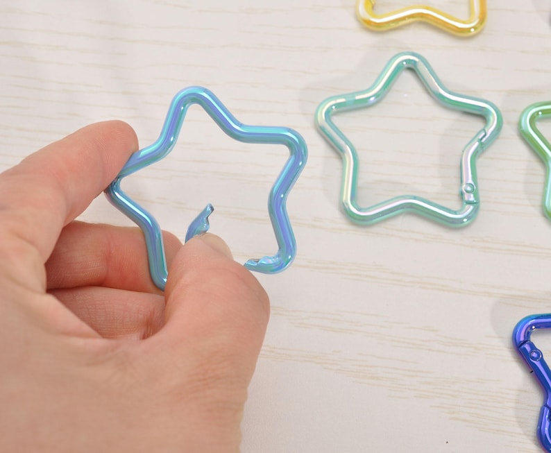 May include: A hand holding a blue star-shaped carabiner clip. Several other star-shaped clips in various colors, including green and gold, are visible in the background. The clips have a metallic, iridescent finish.