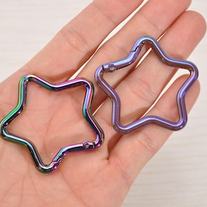 May include: Two star-shaped carabiner clips with a metallic, iridescent finish. One clip is open, and the other is closed. The colors shift from green to purple and blue. These clips are likely used for attaching items.