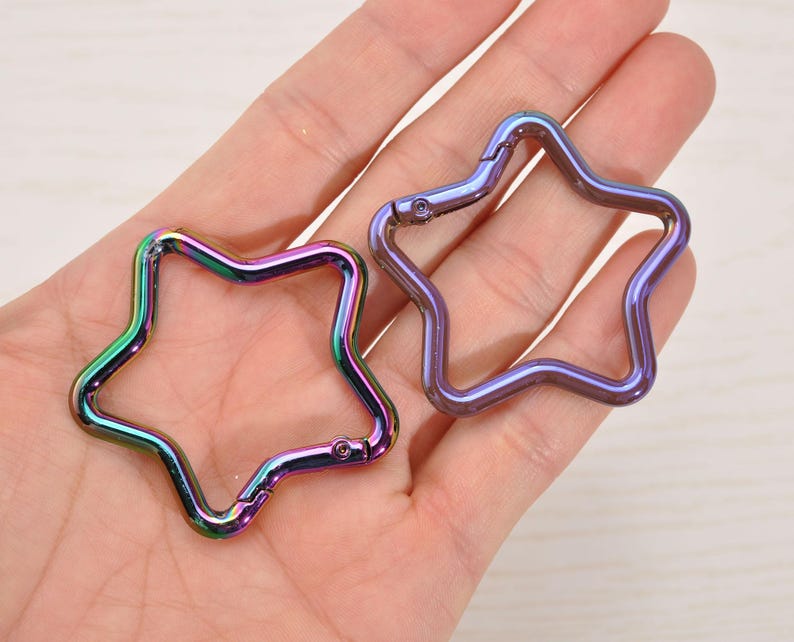 May include: Two star-shaped carabiner clips with a metallic, iridescent finish. One clip is open, and the other is closed. The colors shift from green to purple and blue. These clips are likely used for attaching items.