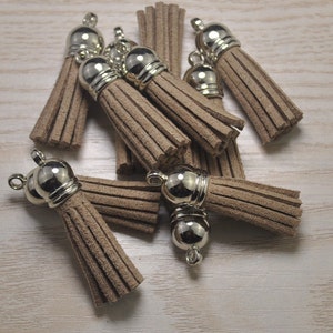 Könnte beinhalten: Eine Sammlung von hellbraunen Wildlederquasten mit silberfarbenen Metallkappen und Schlaufen. Die Quasten sind in einem Haufen angeordnet und zeigen ihre weiche Textur und ihr dekoratives Potenzial. Ideal für die Schmuckherstellung oder Bastelprojekte.