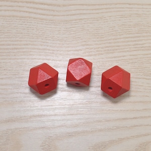 May include: Three red wooden beads with a geometric, faceted design. Each bead has a small hole drilled through the center. The beads are arranged on a light wood grain surface.