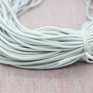 Può includere: Un fascio di cordoncino elastico grigio chiaro, avvolto strettamente e fissato con un cordoncino abbinato. Il cordoncino appare liscio e flessibile, adatto a vari progetti di artigianato e cucito. Lo sfondo è un tessuto rosa tenue.