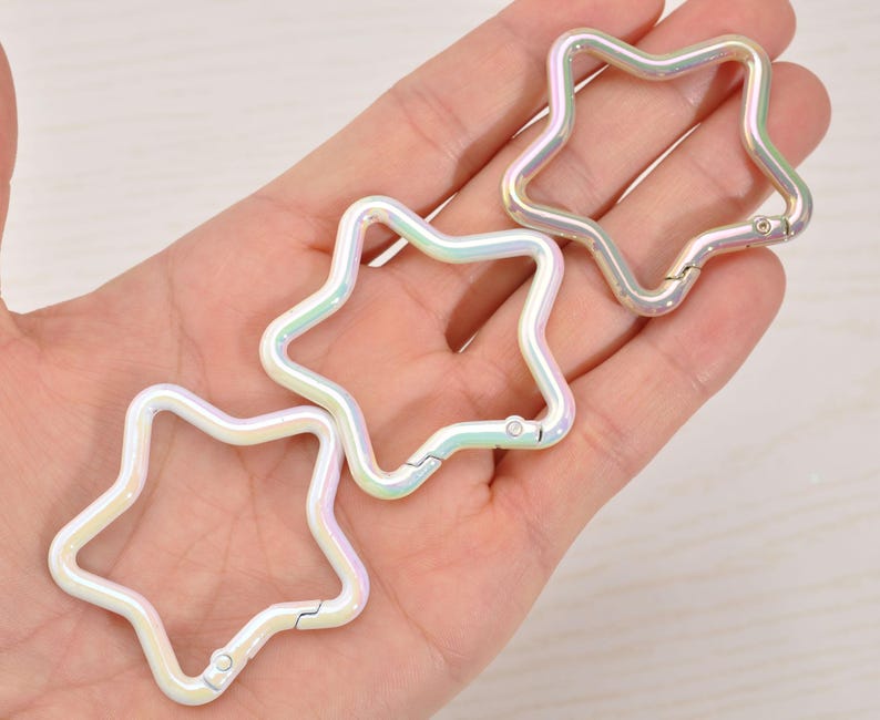 May include: Three iridescent star-shaped clasps. The clasps are made of metal and have a pearlescent finish that reflects a rainbow of colors. Each clasp has a hinged opening for attaching to other items.
