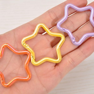 May include: Three star-shaped carabiner clips in orange, yellow, and purple. The clips are made of metal and have a glossy finish. The clips are held in a hand.