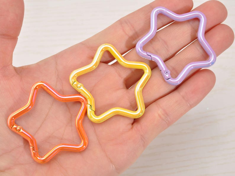 May include: Three star-shaped carabiner clips in orange, yellow, and purple. The clips are made of metal and have a glossy finish. The clips are held in a hand.