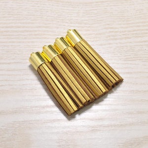 Könnte beinhalten: Fünf gold-braune Lederquasten. Jede Quaste hat eine goldfarbene Metallkappe und eine Reihe dünner, vertikaler Lederstreifen in einem warmen Braunton. Die Quasten sind diagonal auf einer hellen Holzmaserung angeordnet.