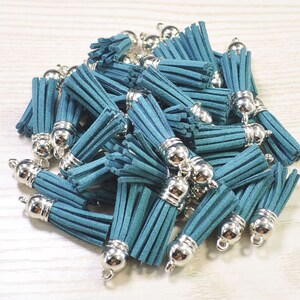 Puede incluir: Un montón de borlas de gamuza azul turquesa con tapas de metal plateado. Las borlas miden aproximadamente 5 cm de largo.