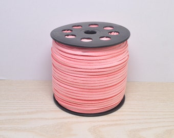 May include: A spool of pink suede cord. The cord is neatly wound around a black plastic spool. The pink cord is a soft, pastel color, and the spool is sitting on a light-colored wooden surface.