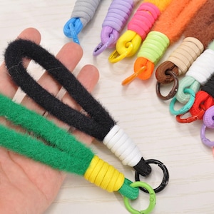 Può includere: Una varietà di cordini da polso colorati. I cordini sono realizzati in morbida corda sfocata in vari colori, tra cui verde, nero, arancione e viola. Ogni cordino ha un anello di metallo per attaccare chiavi o altri oggetti. I cordini sono mostrati in una mano.