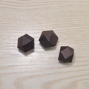 Puede incluir: Tres cuentas de madera marrón oscuro con un diseño geométrico facetado. Cada cuenta tiene un pequeño agujero perforado, lo que sugiere que son para ensartar. Las cuentas se muestran sobre una superficie clara con vetas de madera.