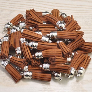 Könnte beinhalten: Eine Ansammlung brauner Wildlederquasten mit silberfarbenen Metallkappen und Schlaufen. Die Quasten sind dicht beieinander, was ihr strukturiertes, gefranstes Design hervorhebt. Diese dekorativen Elemente eignen sich für die Schmuckherstellung, Bastelarbeiten oder zum Verzieren.