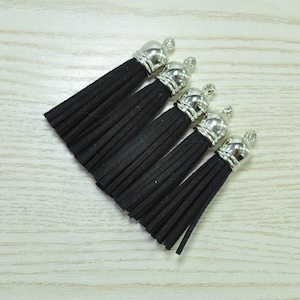 Puede incluir: Cinco borlas de ante negro con tapas de metal plateado. Las borlas están dispuestas en diagonal sobre una superficie clara con vetas de madera. Cada borla tiene un lazo en la parte superior para sujetar.