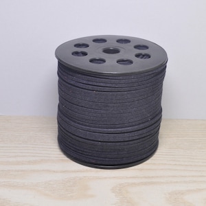May include: A spool of dark gray, flat, round cord. The cord is tightly wound on a black plastic spool with multiple holes. The cord appears to be made of a soft, felt-like material. The spool is sitting on a light-colored wooden surface.