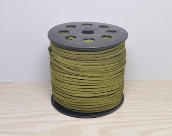 May include: A spool of olive green suede cord. The cord is tightly wound around a black plastic spool with multiple holes on top. The cord appears to be approximately 3mm thick. The spool is sitting on a light wood surface.