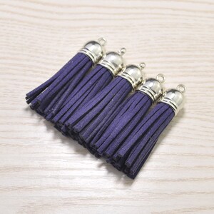 May include: Five dark purple suede tassels with silver-colored metal caps and small loops. The tassels are arranged in a diagonal line on a light wood-grain surface. These are suitable for jewelry making or craft projects.