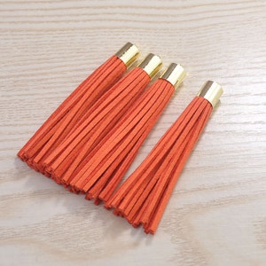 Könnte beinhalten: Vier orangefarbene Lederquasten mit goldfarbenen Metallkappen. Die Quasten sind diagonal auf einer hellen Holzmaserung angeordnet. Der Fransenrand jeder Quaste ist sichtbar, und die Farbe ist ein leuchtendes, warmes Orange.