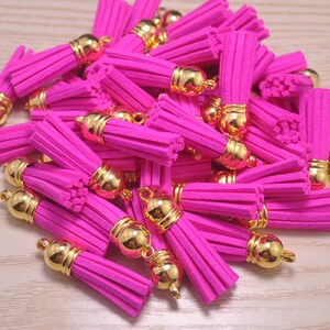 Könnte beinhalten: Ein Haufen leuchtend pinkfarbener Quasten mit goldfarbenen Kappen und Schlaufen. Die Quasten bestehen aus einem weichen, Wildleder-ähnlichen Material. Die goldenen Kappen haben eine abgerundete Form und sind am Quastenkörper befestigt.