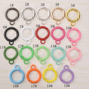 May include: A collection of nineteen round metal clasp keychains in various colors, including black, silver, gold, red, pink, green, blue, orange, yellow, and purple. Each keychain has a spring-loaded clasp and a small loop.
