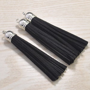 May include: Close-up of three black tassels with silver-colored metal caps. The tassels are made of a soft, textured material and are arranged on a light-colored wooden surface. The tassels are a craft supply.